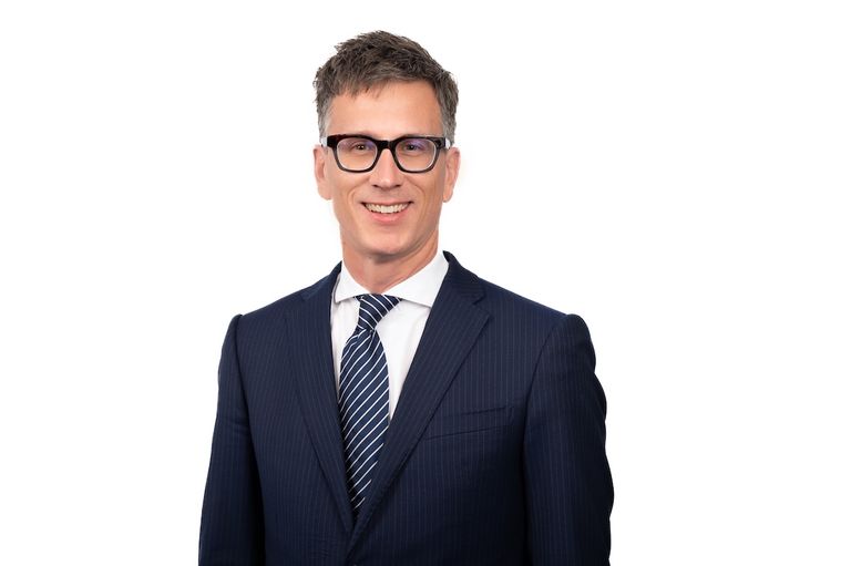 Professional headshot of Adrian Tisato wearing glasses and a navy suit against a white background.
